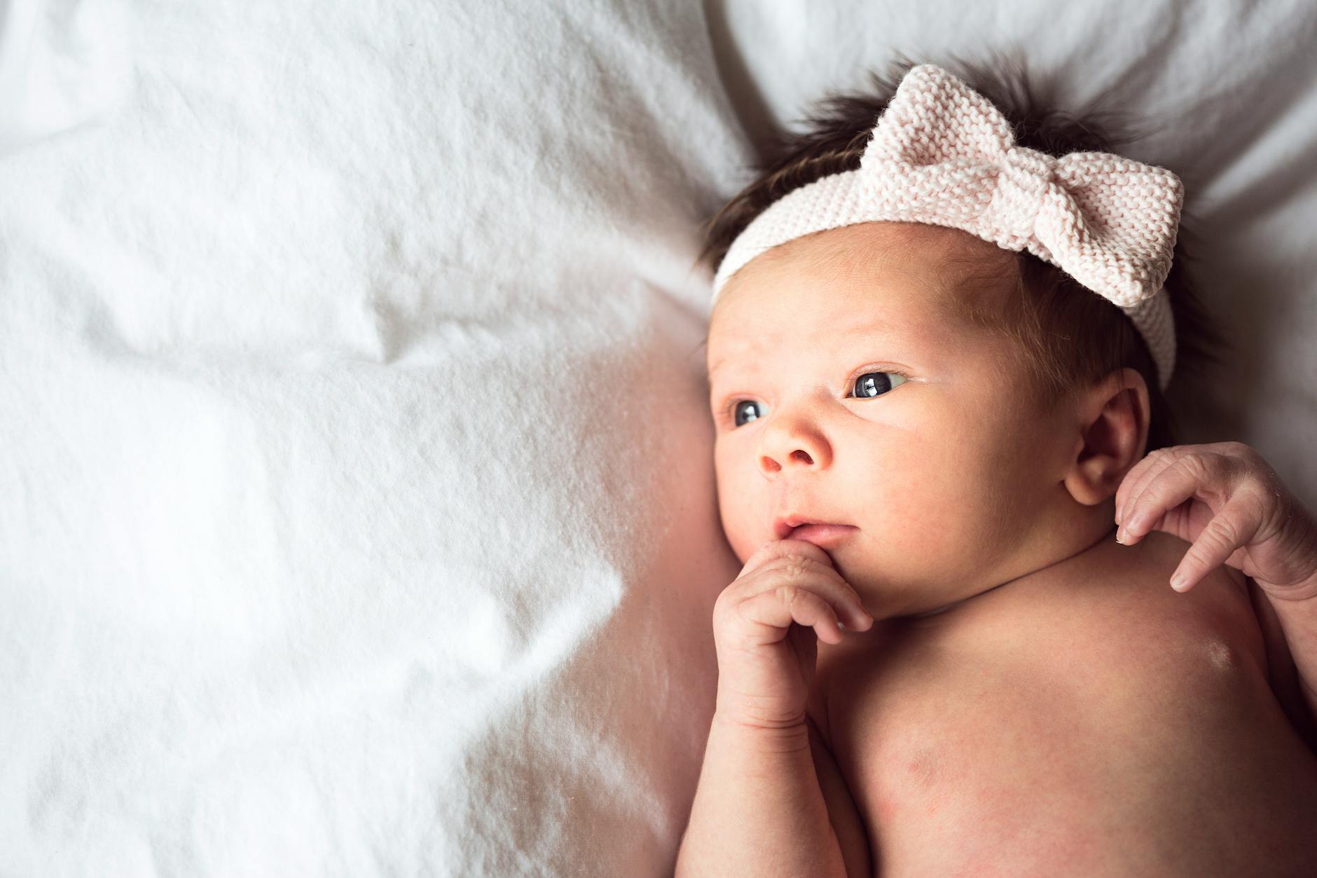 Ilustrasi: Charming portrait of a newborn baby with a pink bow headband lying on a soft white blanket.