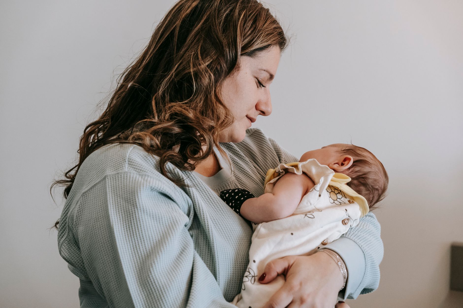 Ilustrasi: Side view of caring mother with sleeping newborn baby in arms standing on white background in light room at home