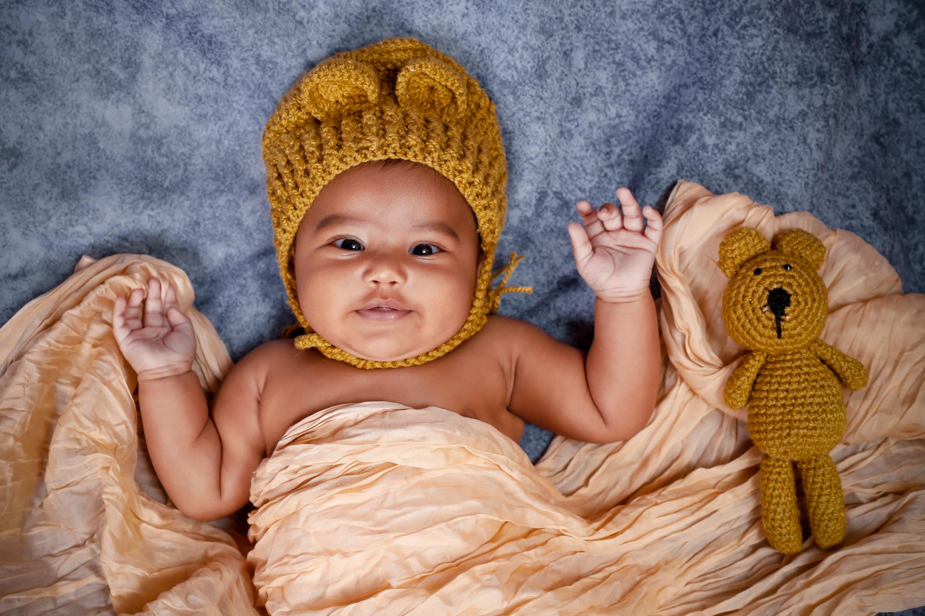 Ilustrasi: Charming newborn baby with a knitted cap lying on a blanket beside a crochet teddy bear.
