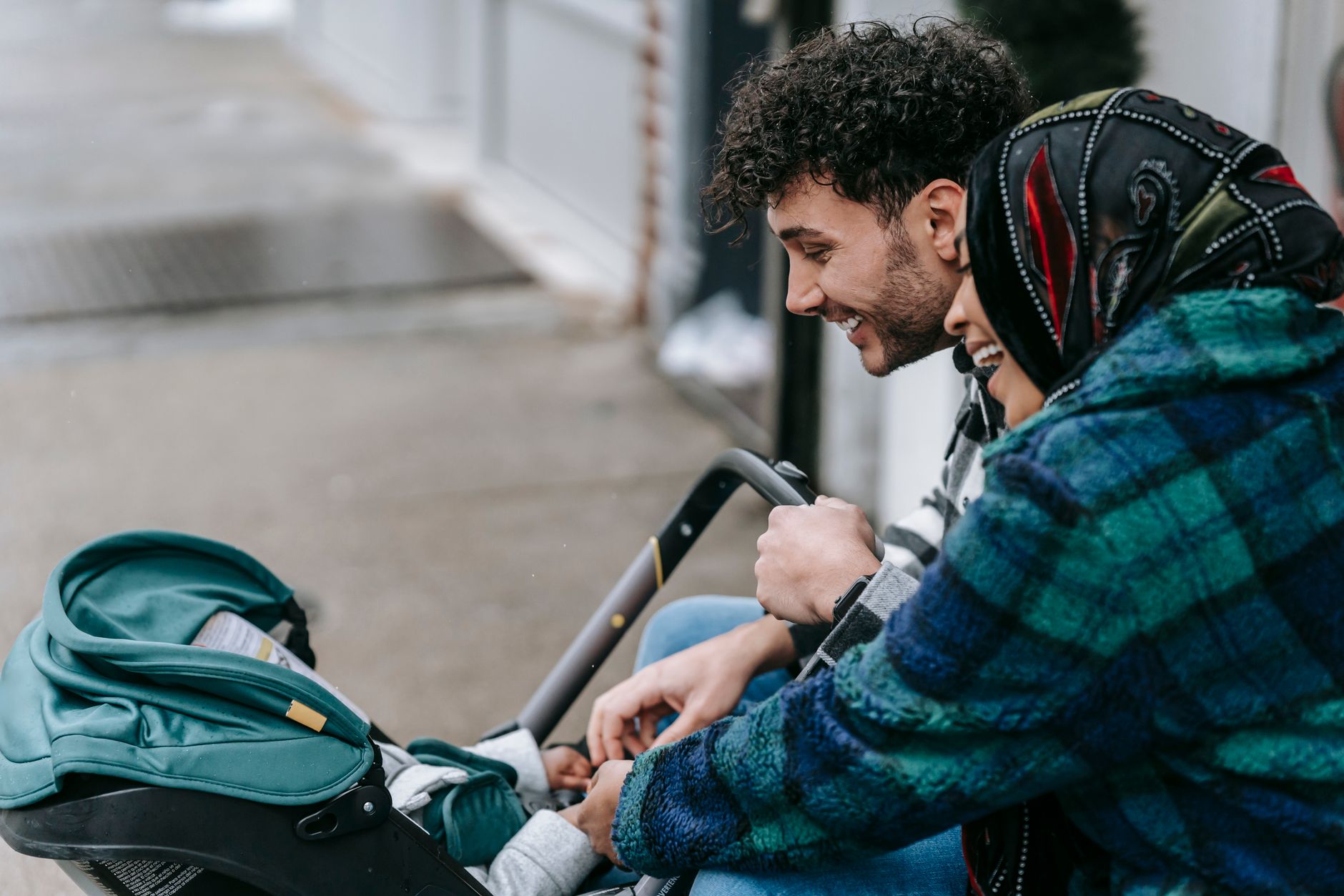 Ilustrasi: Side view of smiling loving ethnic couple holding hands of little baby infant lying in stroller