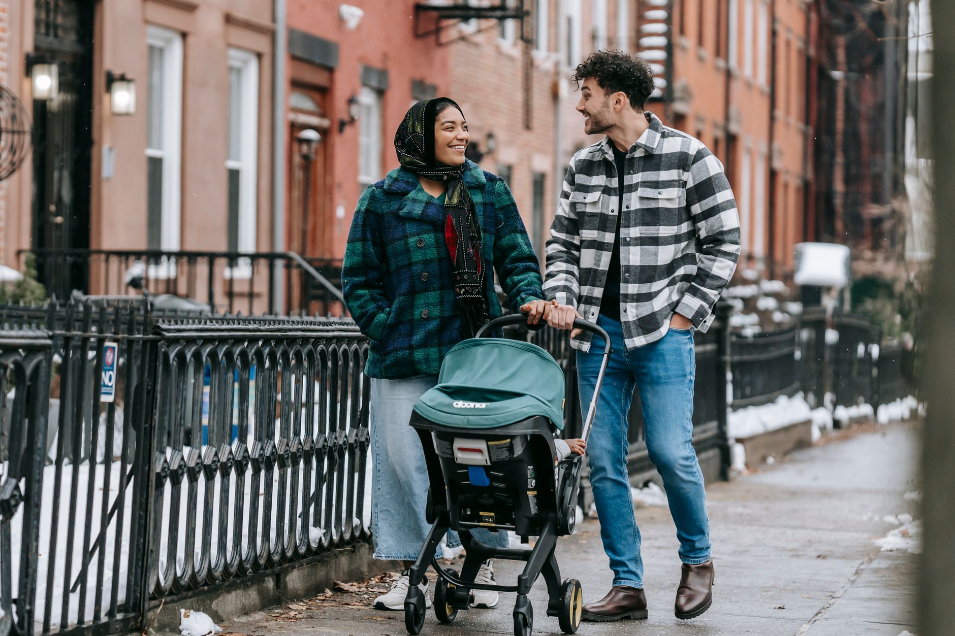Ilustrasi: Full body of multiracial parents looking at each other while strolling on sidewalk along buildings with anonymous child in baby carriage