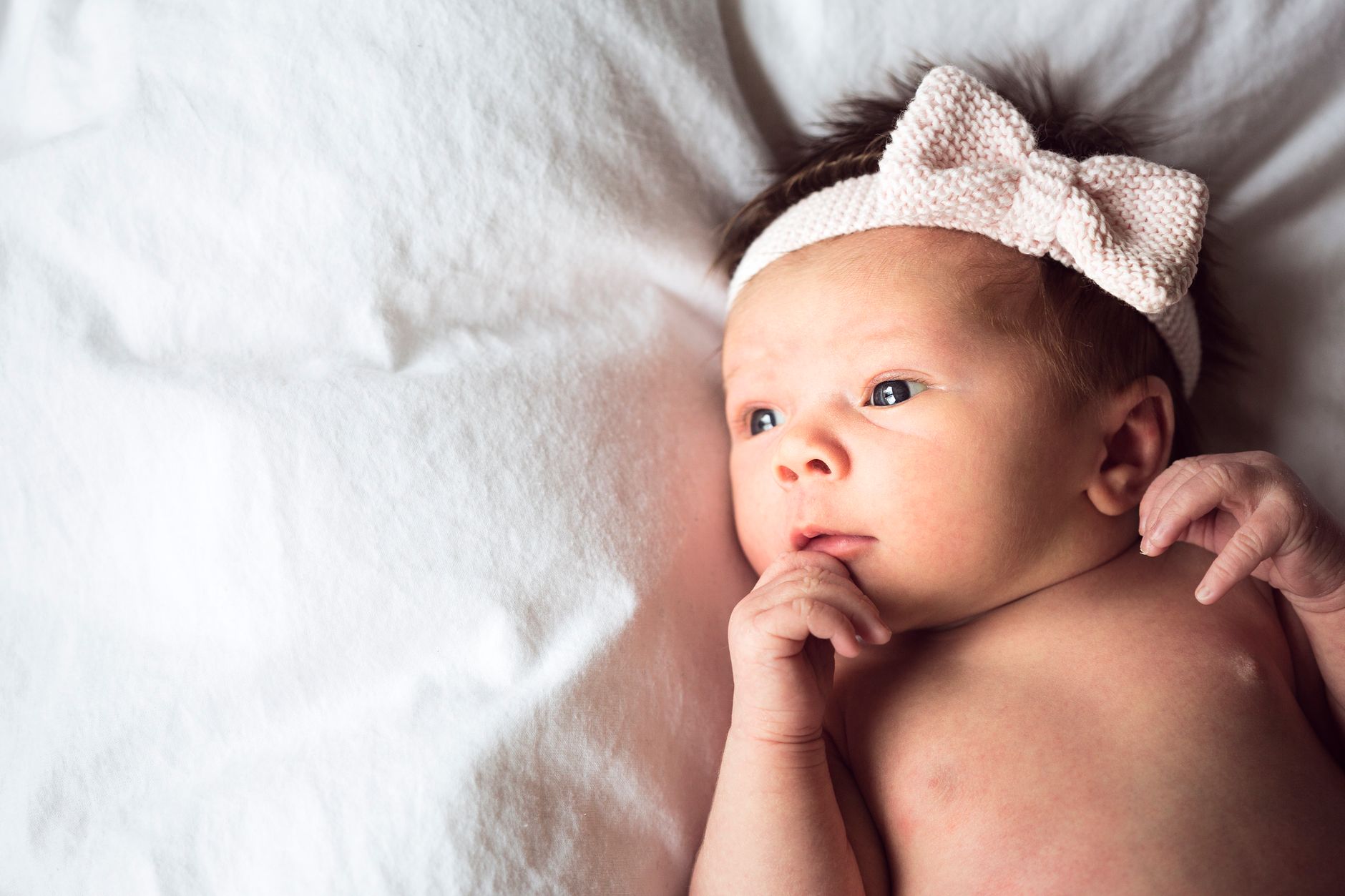 Ilustrasi: Charming portrait of a newborn baby with a pink bow headband lying on a soft white blanket.
