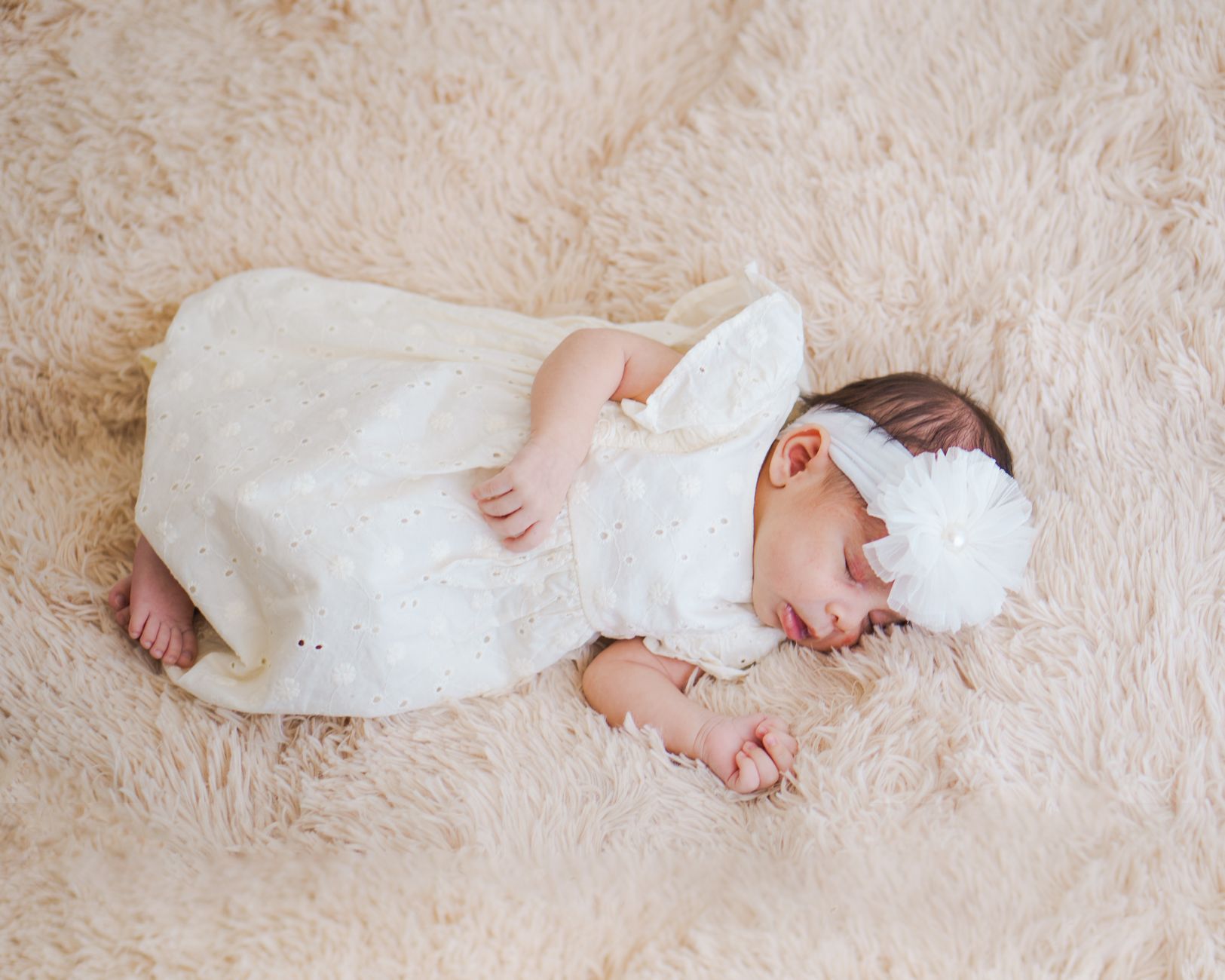 Ilustrasi: Newborn baby girl peacefully sleeping on a fluffy rug wearing a white dress and headband.