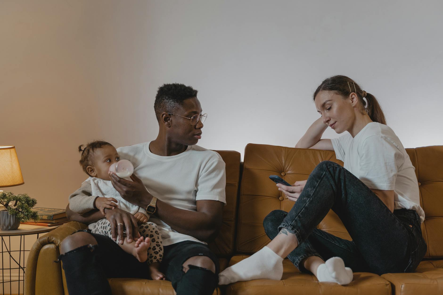 Ilustrasi: Parents relaxing on a sofa while feeding their baby, enjoying family time with a cellphone.