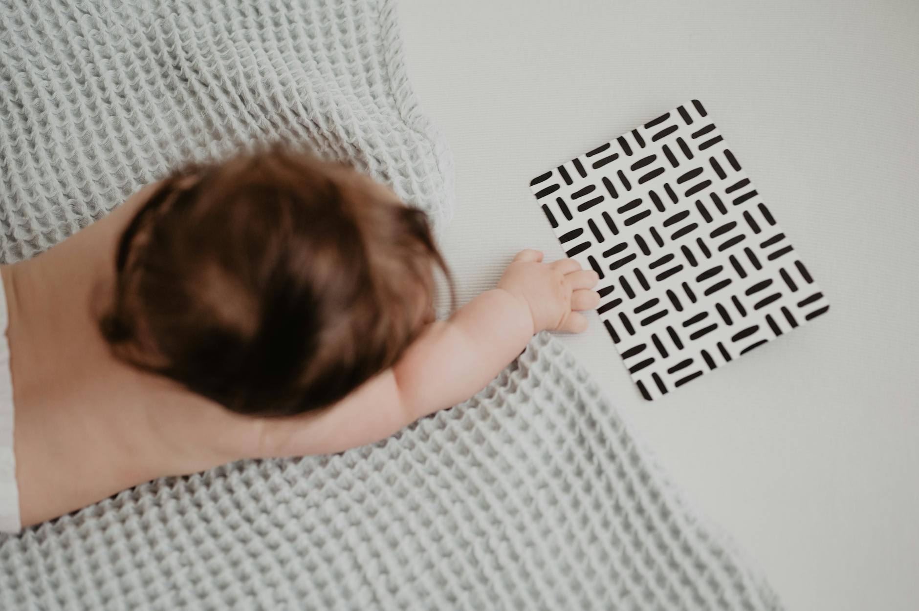 Ilustrasi: An adorable baby lying on a blanket reaching for a geometric patterned card, symbolizing curiosity and exploration.