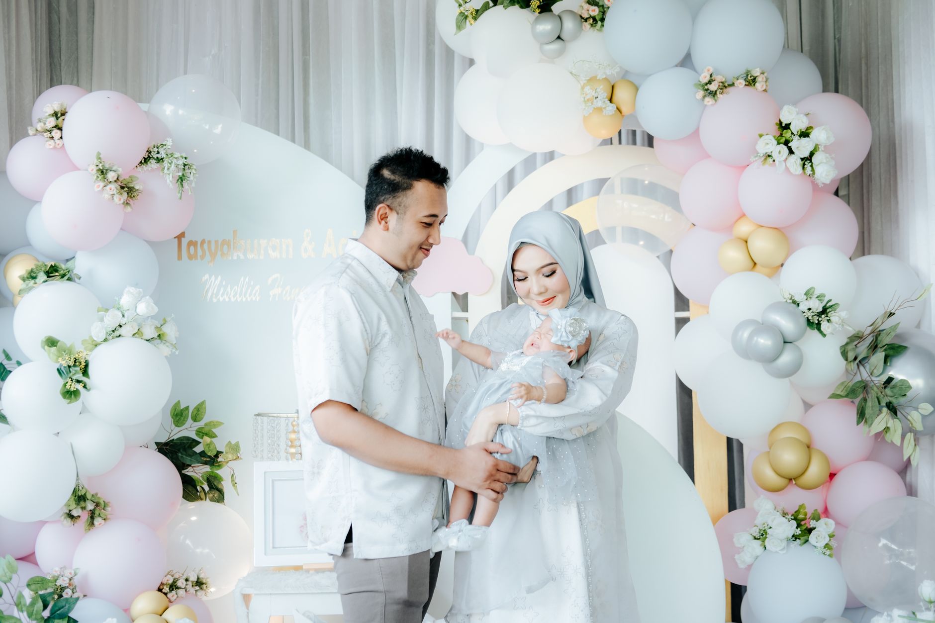 Ilustrasi: A family celebrating with pastel balloons, parents in elegant attire holding their baby.