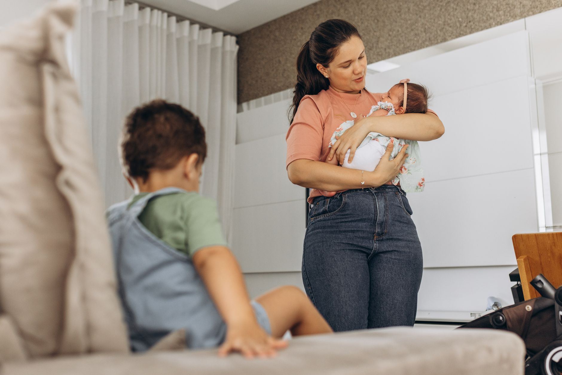 Ilustrasi: A mother lovingly cradles her baby while her child sits nearby on the couch in a modern room setting.