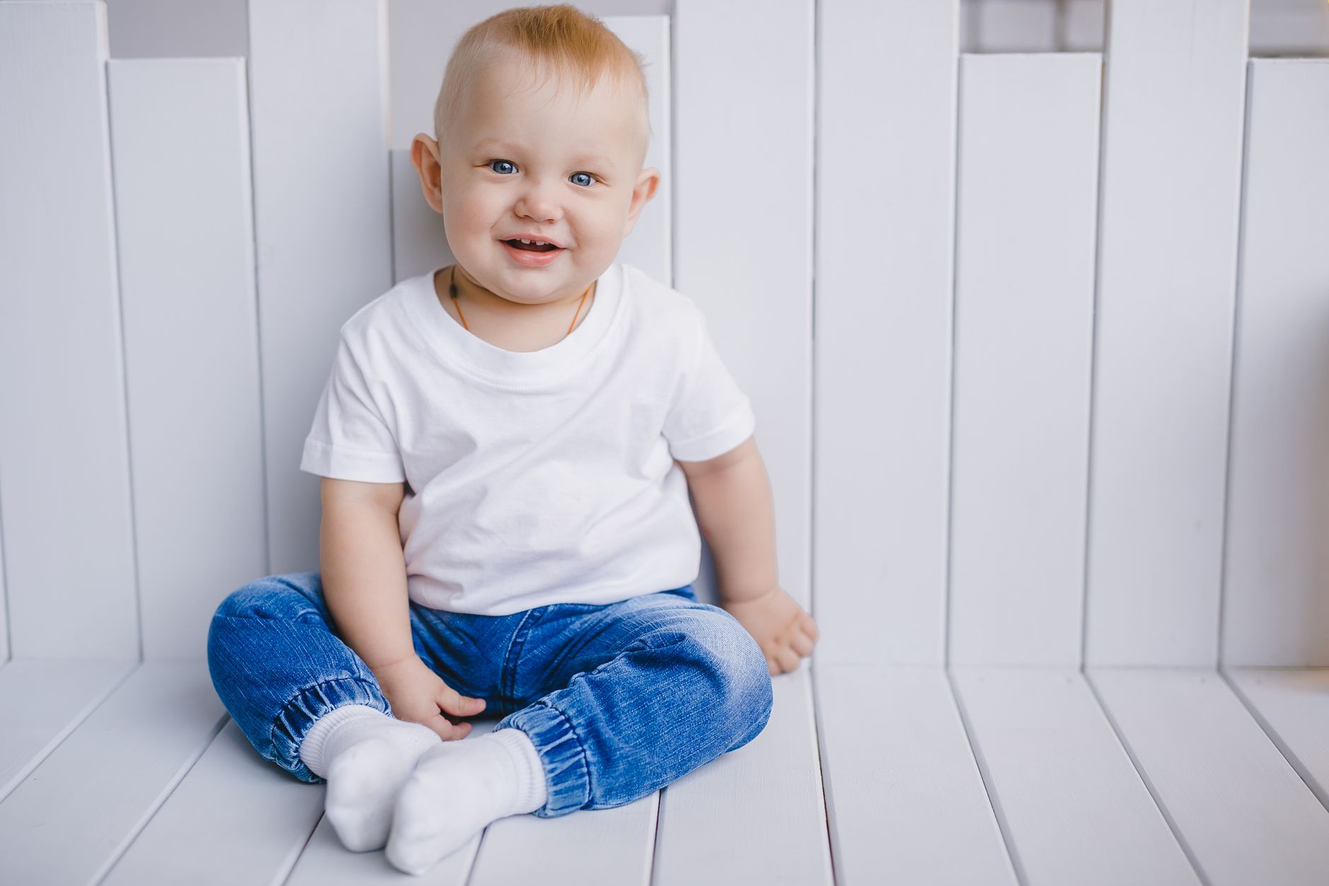 Ilustrasi: Charming portrait of a smiling baby boy in a white t-shirt and blue jeans, sitting indoors.