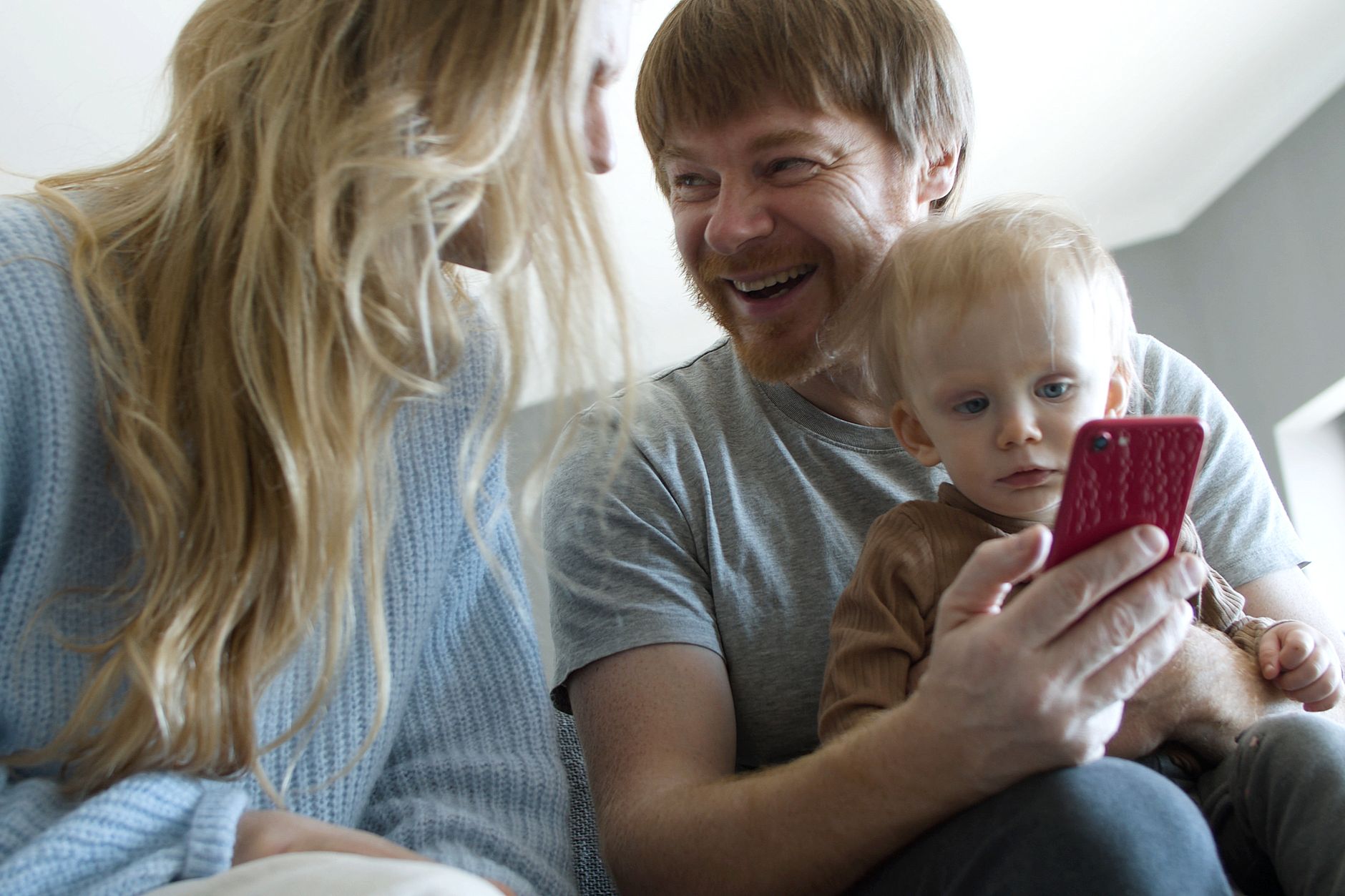 Ilustrasi: A happy family with a baby boy sharing a moment while looking at a smartphone indoors.