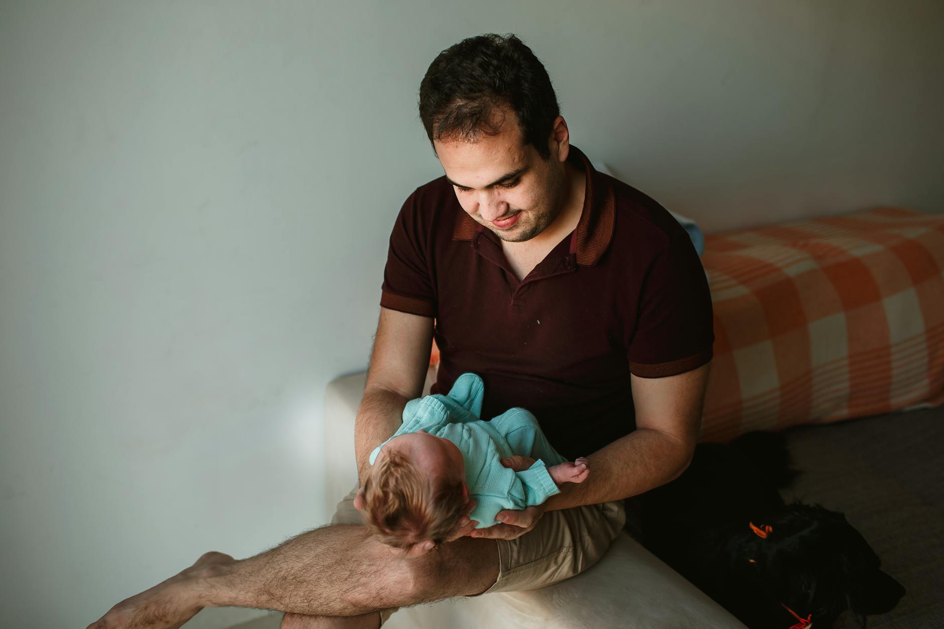 Ilustrasi: Caring father in casual clothes with adorable newborn baby on hands sitting on armrest of couch in light room at home