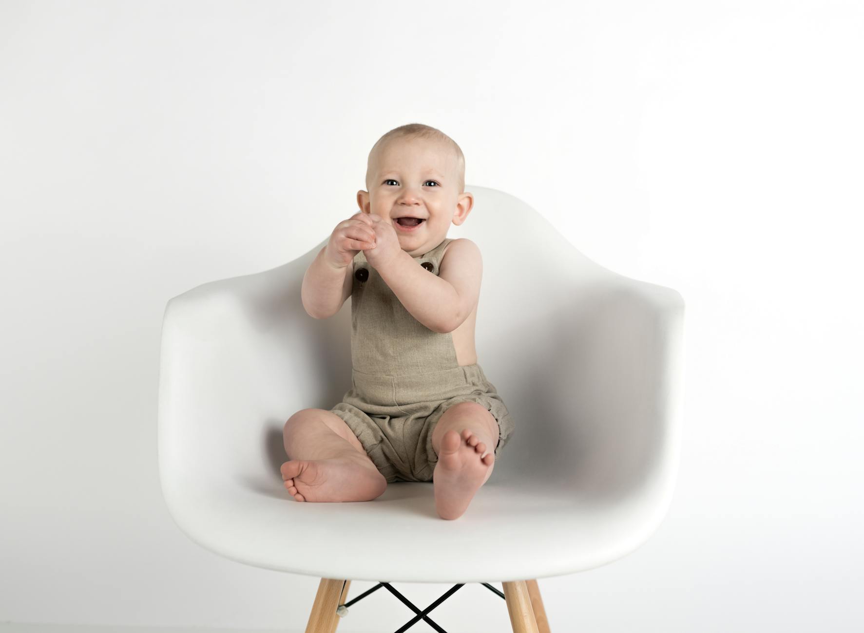 Ilustrasi: Adorable baby boy in beige romper joyfully sitting on a modern white chair indoors.