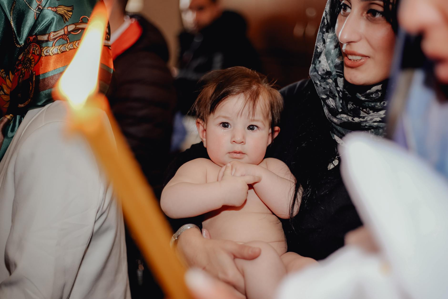 Ilustrasi: A baby held by a mother surrounded by people in a traditional ceremony with a candle.