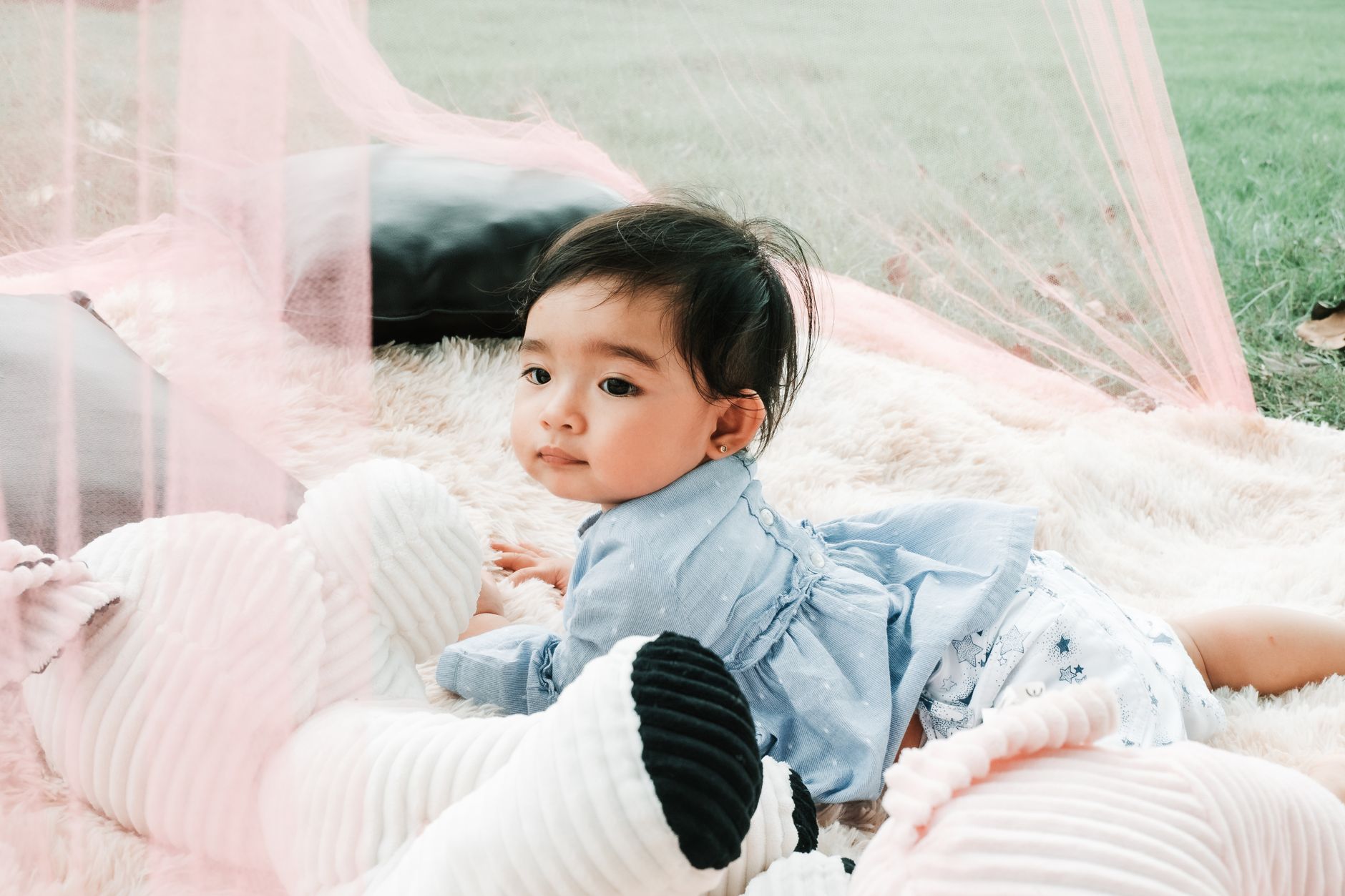 Ilustrasi: Cute baby girl lies on a fluffy rug outside, bermain with a plush toy, surrounded by soft hues.