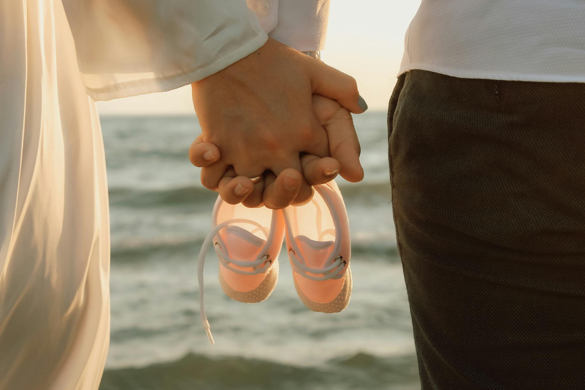Ilustrasi: A couple holding hands and pink baby shoes, standing by the sea during a serene sunset.