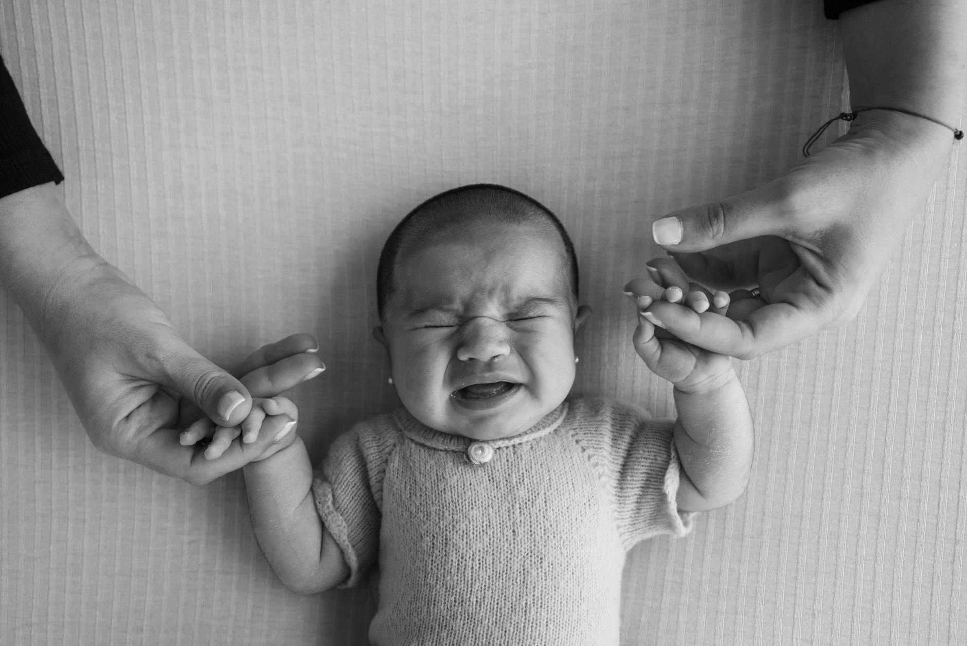 Ilustrasi: Black and white image of a crying baby being comforted by two adult hands holding theirs.