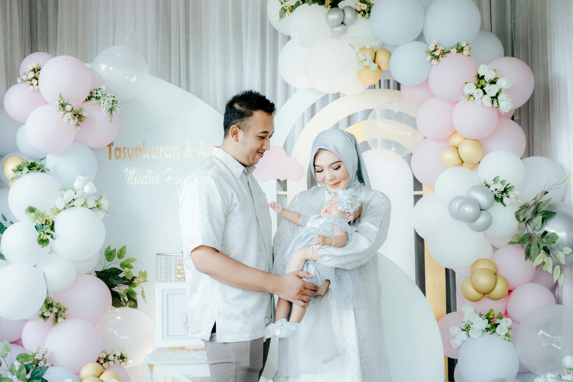 Ilustrasi: A family celebrating with pastel balloons, parents in elegant attire holding their baby.
