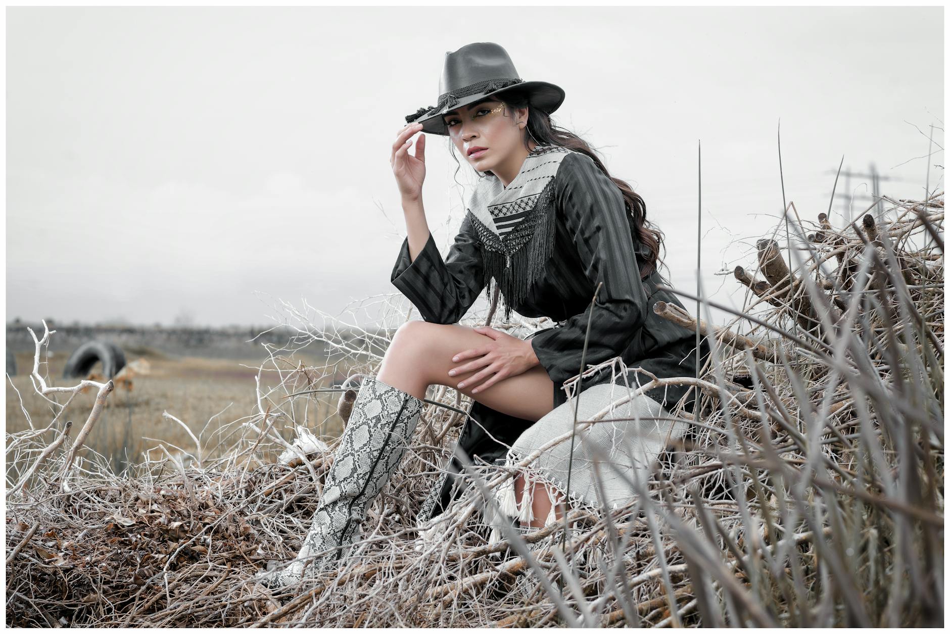 Ilustrasi: A stylish woman in Western attire poses outdoors in a rural setting with dry grass and hay.