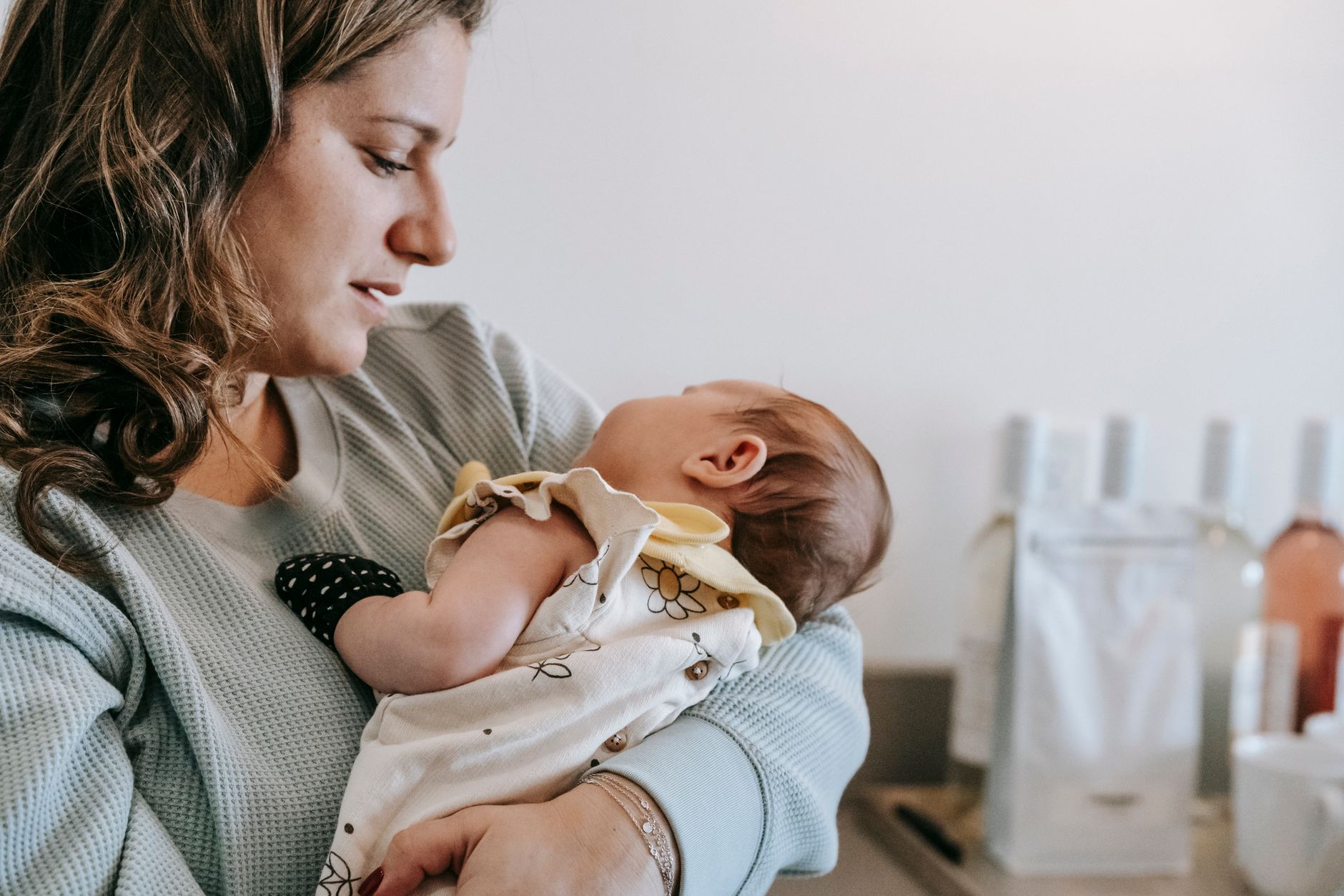 Ilustrasi: Side view of crop caring mother in casual wear with cute newborn baby in arms standing in light room at home