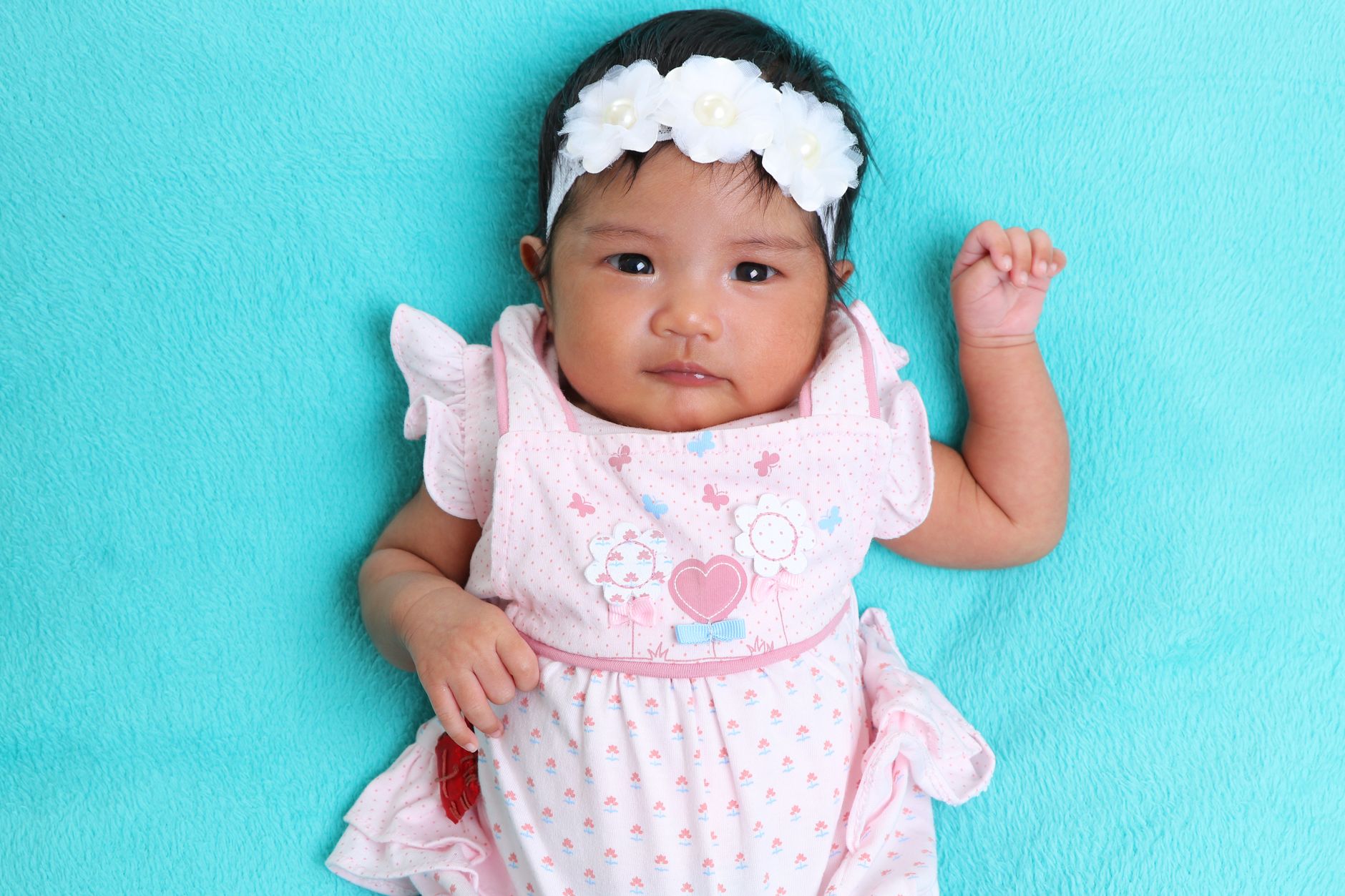 Ilustrasi: Cute baby girl wearing a pink dress and white floral headband, lying on a teal blanket, showcasing innocence.
