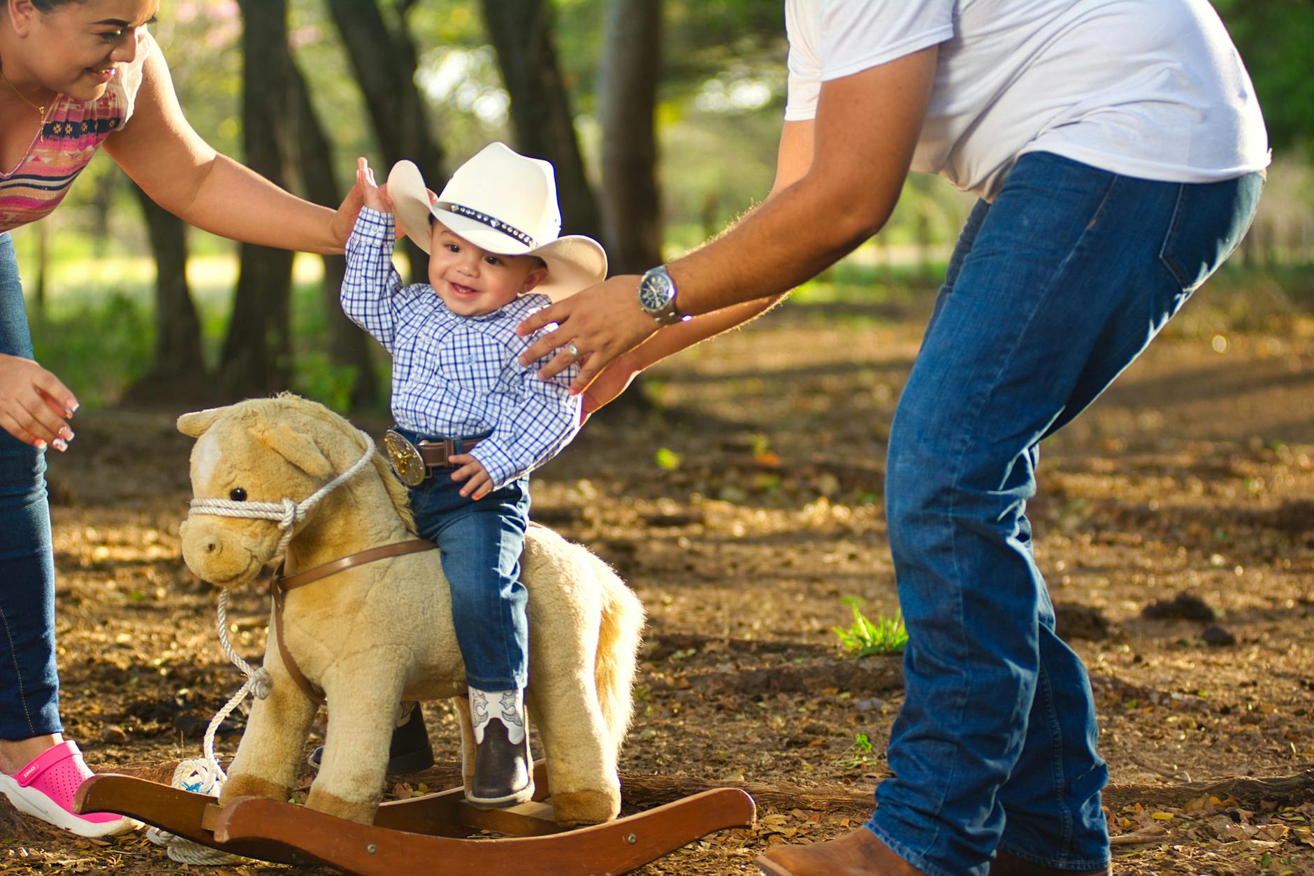 Ilustrasi: Happy baby boy in cowboy hat on plush horse, assisted by parents in park.