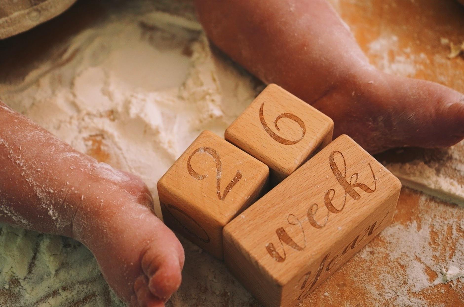 Ilustrasi: Adorable baby feet on flour-covered surface, next to wooden milestone blocks displaying 26 weeks.
