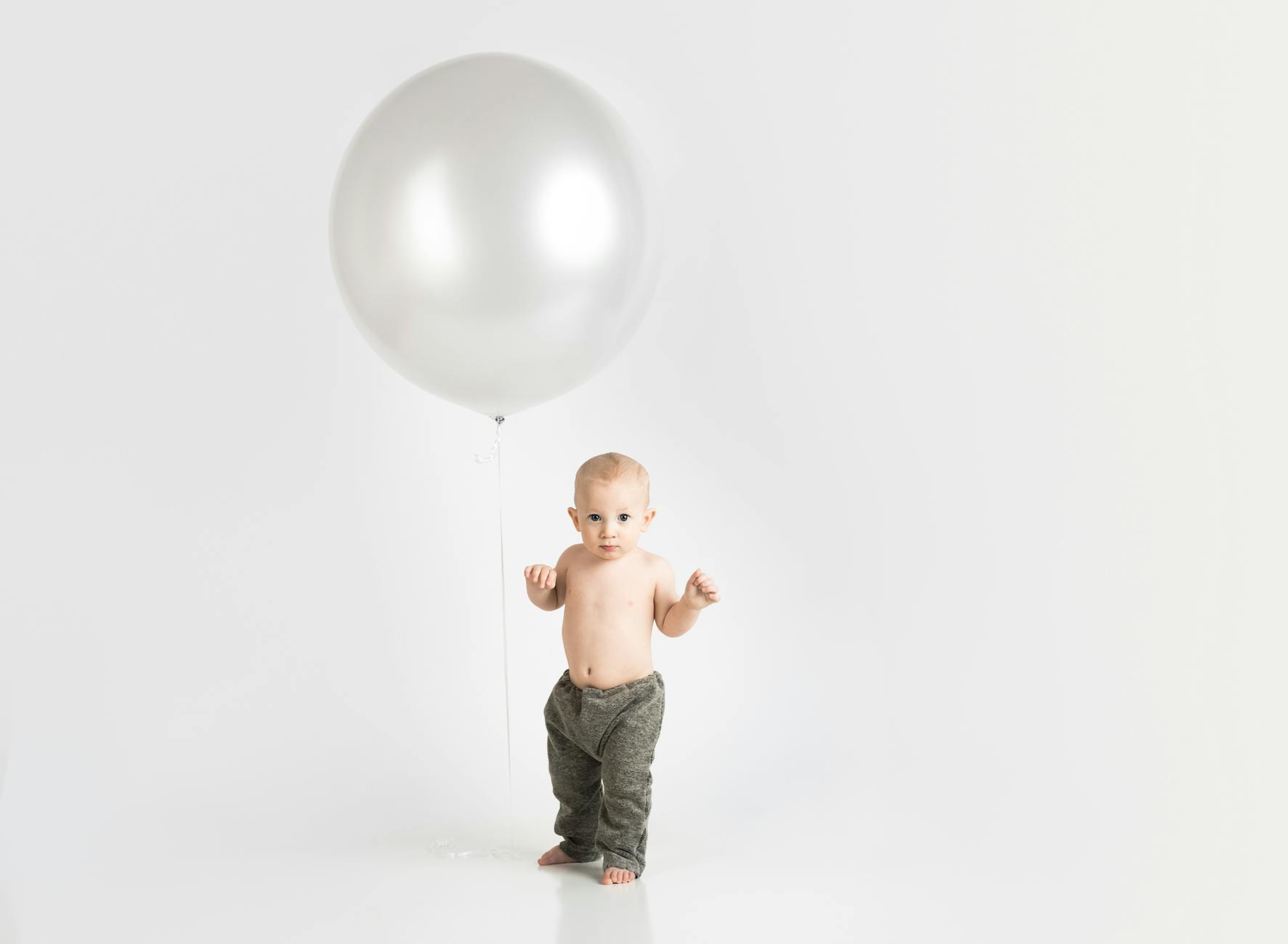Ilustrasi: Cute baby boy standing with a large silver balloon in a bright studio setting.
